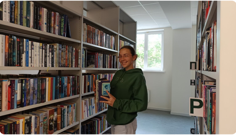 Laura bibliotēkā stāv starp grāmatu plauktiem un rokās tur vairākas grāmatas