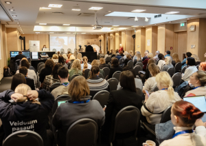 Konferences “Brīvprātīgais darbs Latvijā — vakar, šodien un rīt” foto ar plašu auditoriju, skatuvi un runātāju.