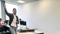 Foto. Uzņēmuma “Trans Speed” pārstāvis birojā stāv pie darba galda un ar paceltu roku skaidro darba procesu; fonā redzami biroja galdi - uzņēmuma darbinieki strādā pie datoriem.