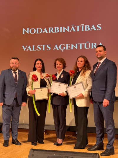 Pieci smaidīgi cilvēki svinīgā pasākumā uz skatuves.