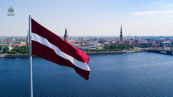 Liels Latvijas valsts karogs, kas plīvo vējā pie Daugavas. Fonā Rīgas panorāma ar Vecrīgas torņiem un pilsētas ēkām saulainā laikā.