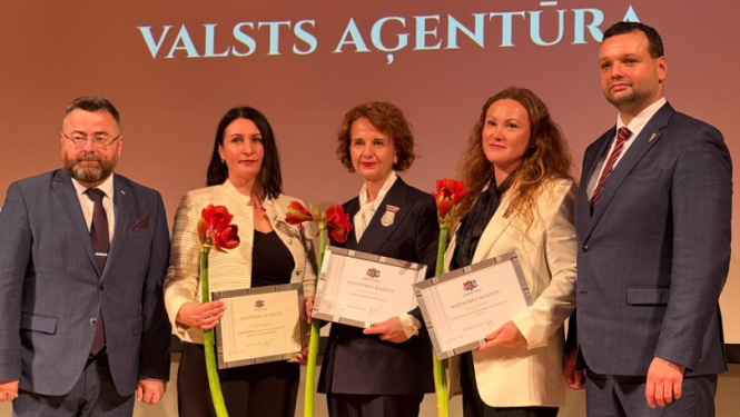 Pieci smaidīgi cilvēki svinīgā pasākumā uz skatuves.