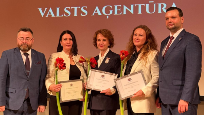 Pieci smaidīgi cilvēki svinīgā pasākumā uz skatuves.