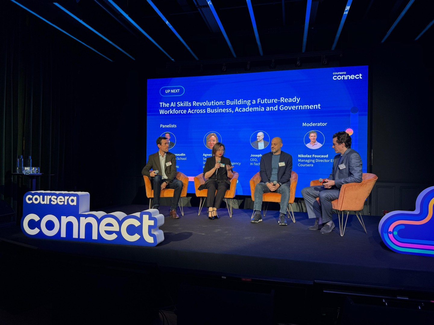 Fotogrāfija no paneļdiskusijas Coursera Connect pasākumā. Uz skatuves redzami četri diskusijas dalībnieki.