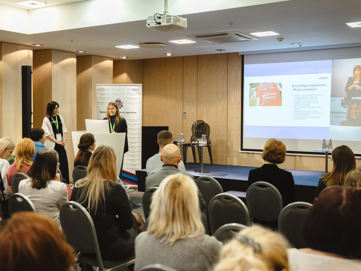 Fotomirkļis ar auditoriju, skatuvi un runātājiem.