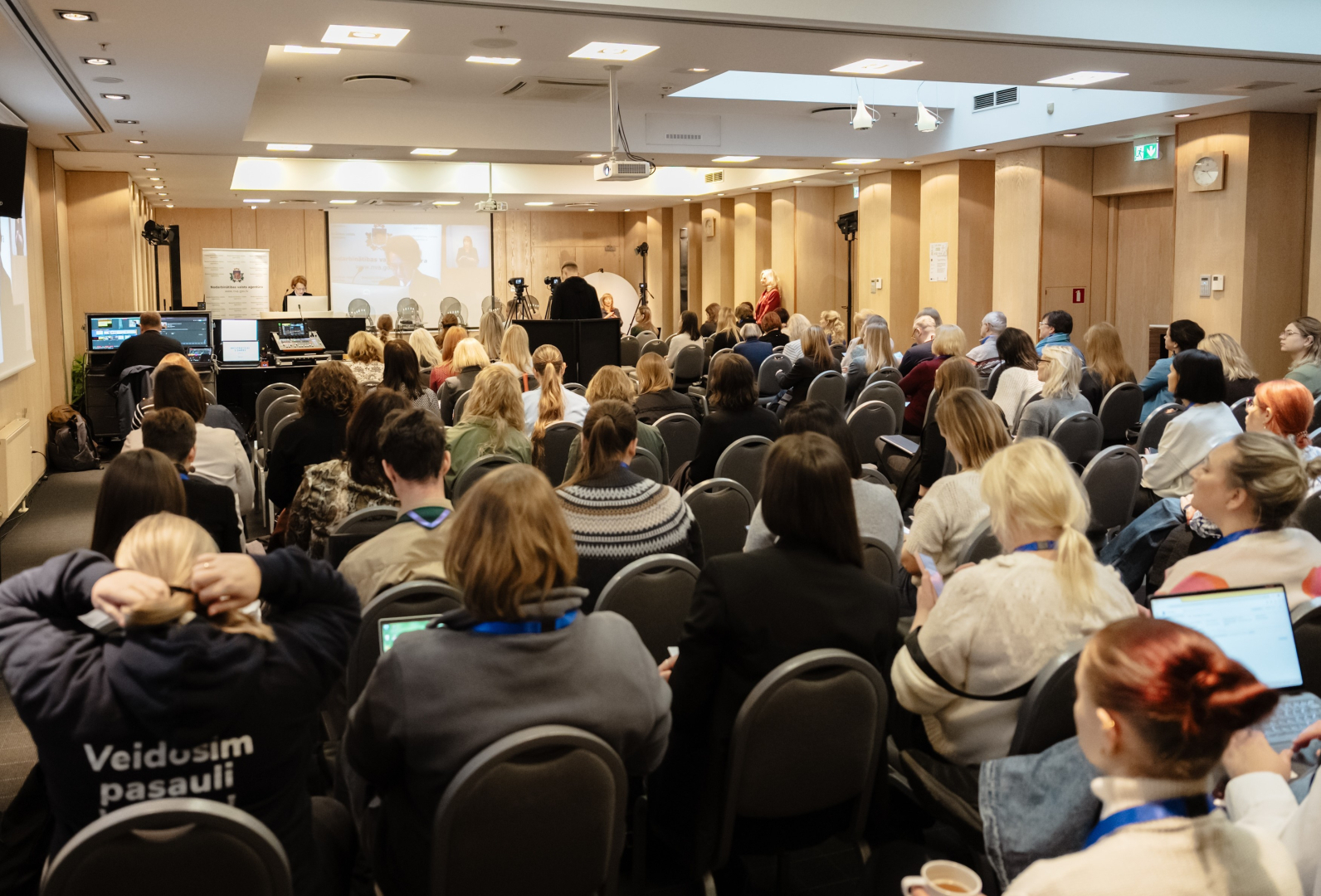 Konferences “Brīvprātīgais darbs Latvijā — vakar, šodien un rīt” foto ar plašu auditoriju, skatuvi un runātāju.