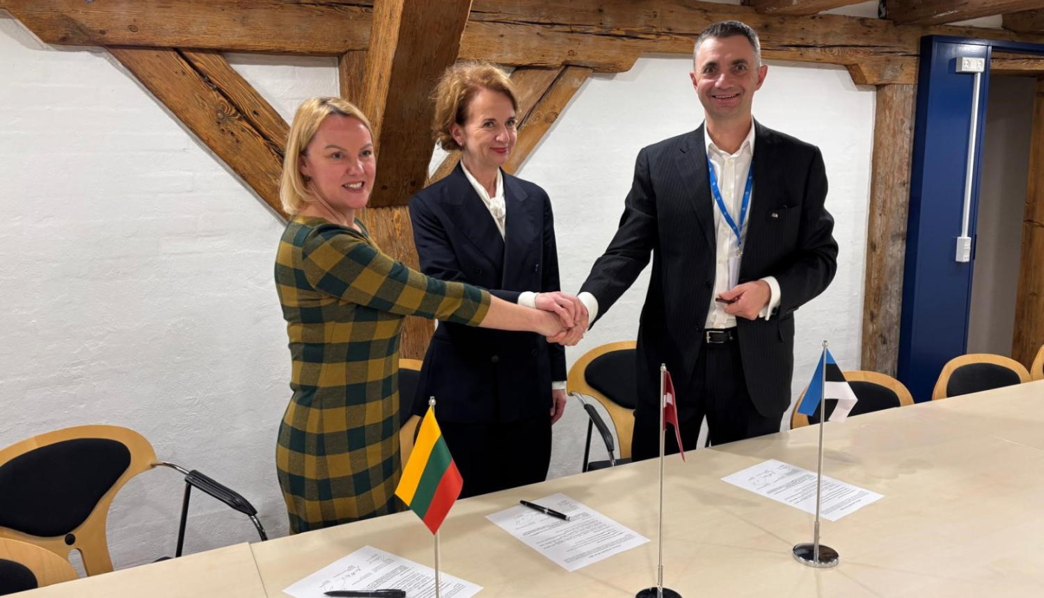 Trīs cilvēki sēž pie galda un paraksta dokumentus; priekšplānā redzami Lietuvas, Latvijas un Igaunijas karodziņi.